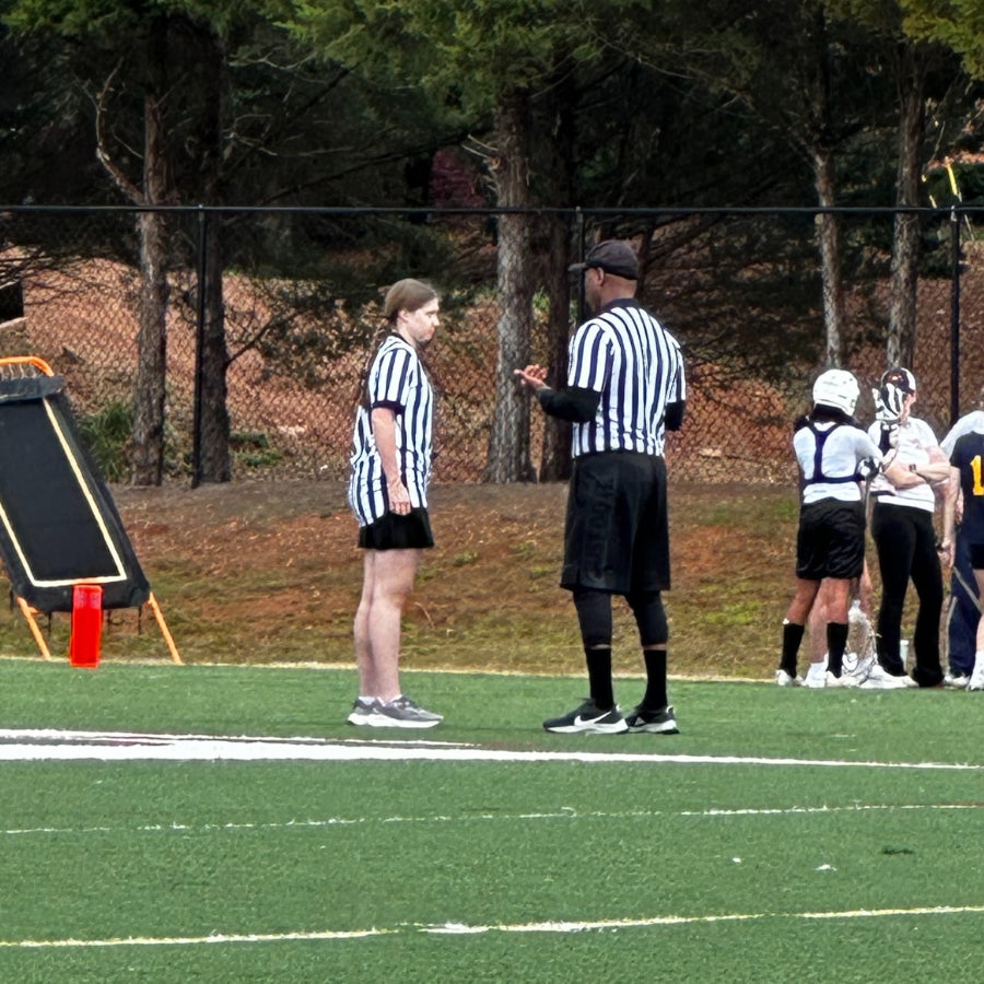 wo lacrosse referees, one a teenage official and one experienced, talk on the field during a youth game — a reminder of respect and mentorship in youth sports.